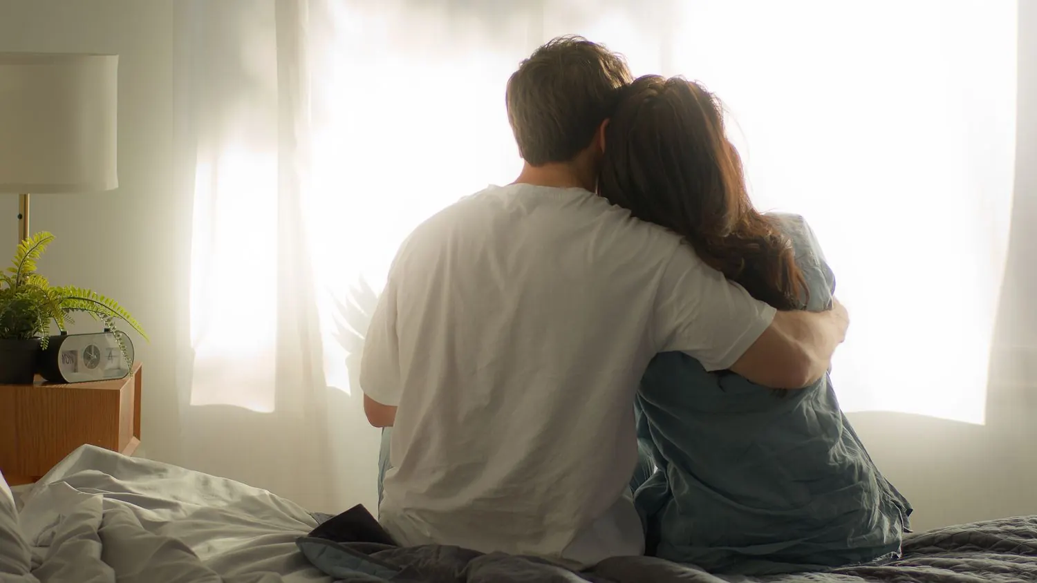A couple sitting on the edge of their bed in morning light, holding each other