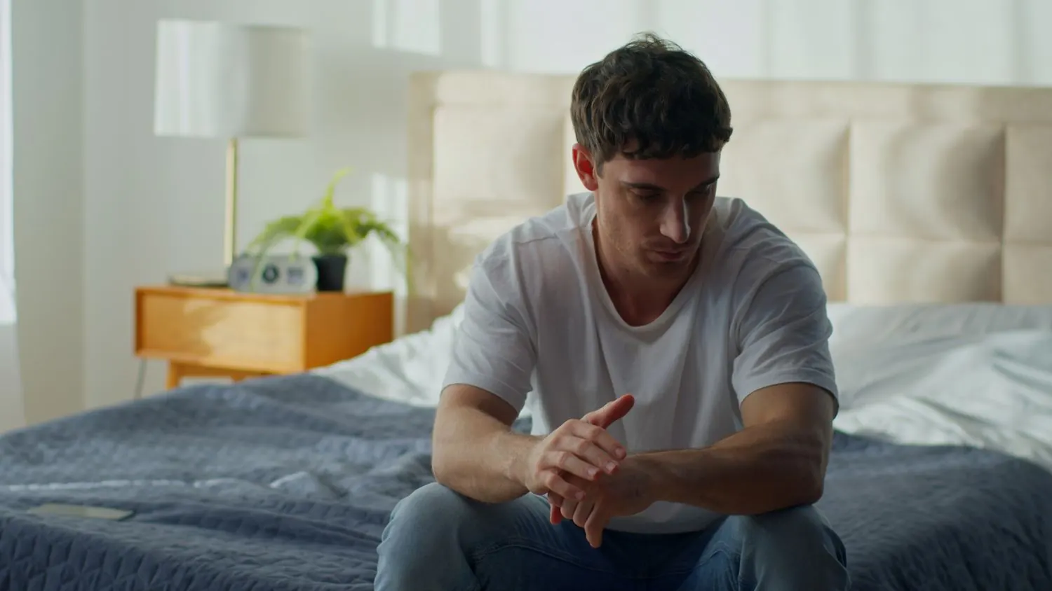 A young man sits on the edge of his bed, head lowered