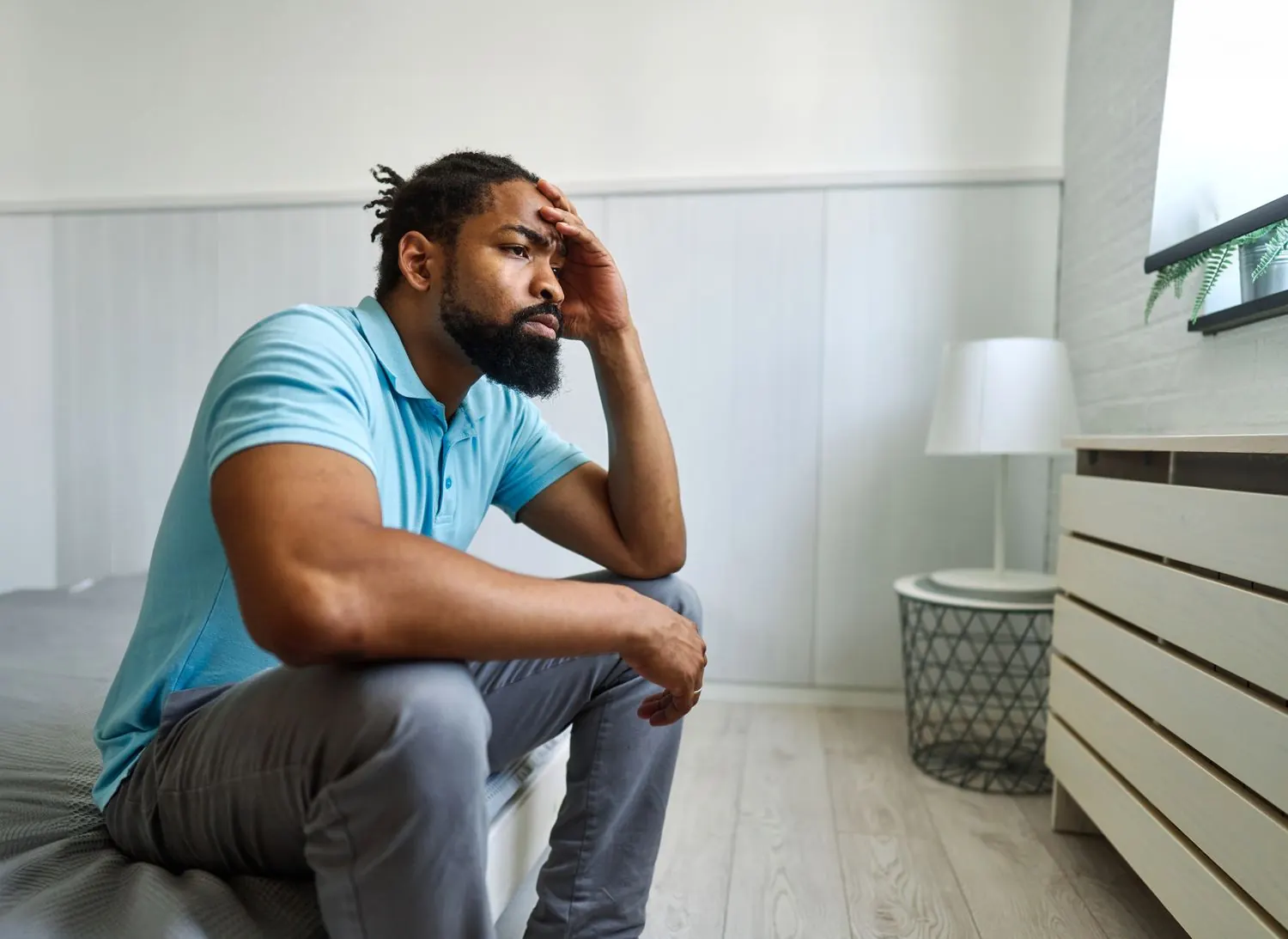 A man sits on his bed with his hand pressed to his forehead